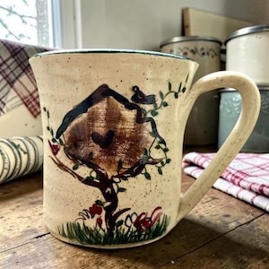 May include: A beige ceramic mug with a hand-painted birdhouse design. The birdhouse is brown with a dark blue roof, and a small bird sits on top. Green vines and red flowers decorate the tree and base of the mug.
