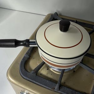 May include: A cream-colored saucepan with a matching lid, featuring a dark brown circular accent on the lid and horizontal brown and black stripes around the body. The pan has a long, dark brown wooden handle and a dark brown knob on the lid.