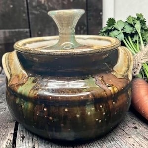May include: A round, glazed ceramic pot with two side handles and a matching lid featuring a central knob. The exterior displays a mottled, drip-style glaze in shades of deep brown, earthy green, and tan, with small white speckles throughout.