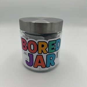 May include: A clear glass jar with a silver lid, filled with small white slips of paper. The jar has a colourful label that reads "BORED JAR" in a playful font. The jar is sitting on a white surface.