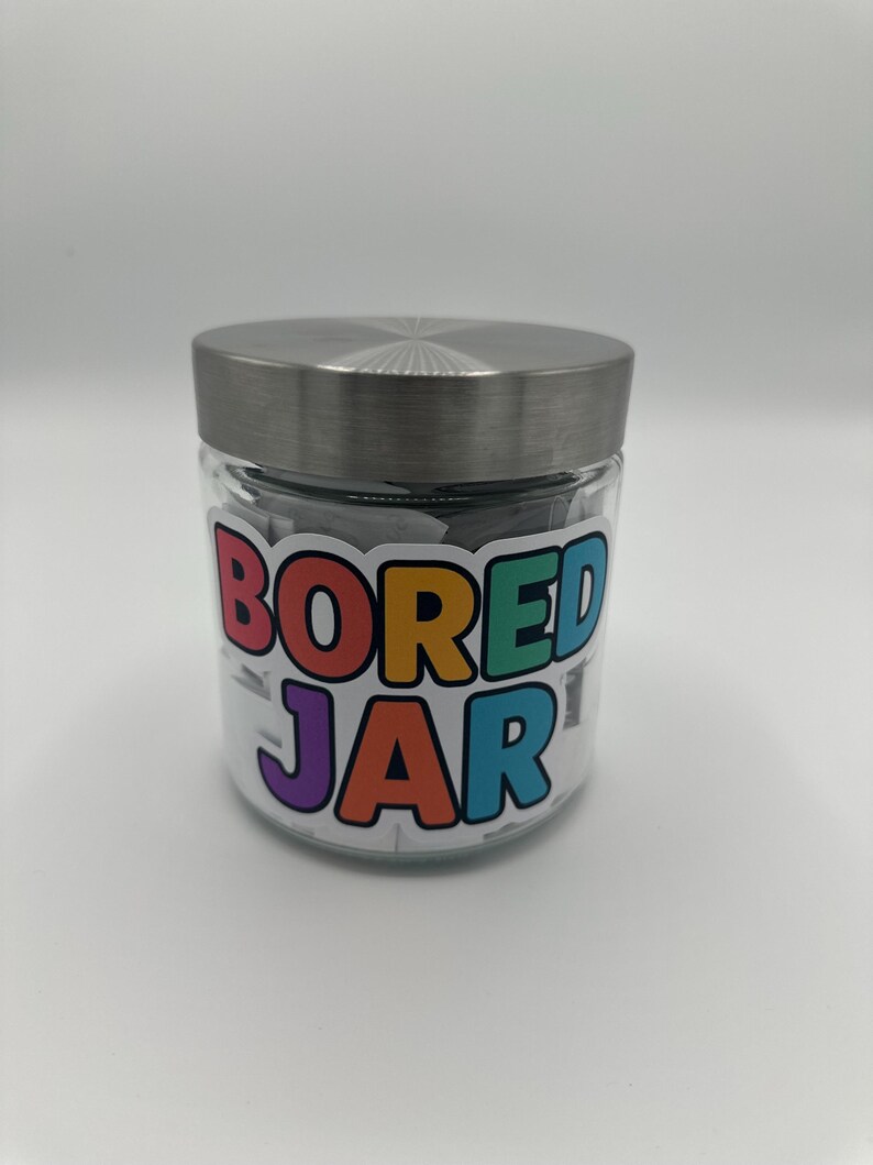 May include: A clear glass jar with a silver lid, filled with small white slips of paper. The jar has a colourful label that reads "BORED JAR" in a playful font. The jar is sitting on a white surface.