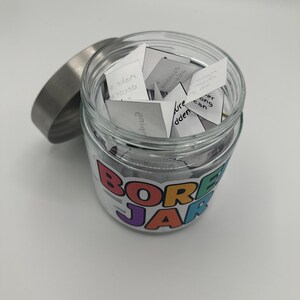 May include: A clear glass jar with a silver lid, filled with small white paper slips. The jar has a colorful label that reads "BORED JAR". The jar is sitting on a white surface.