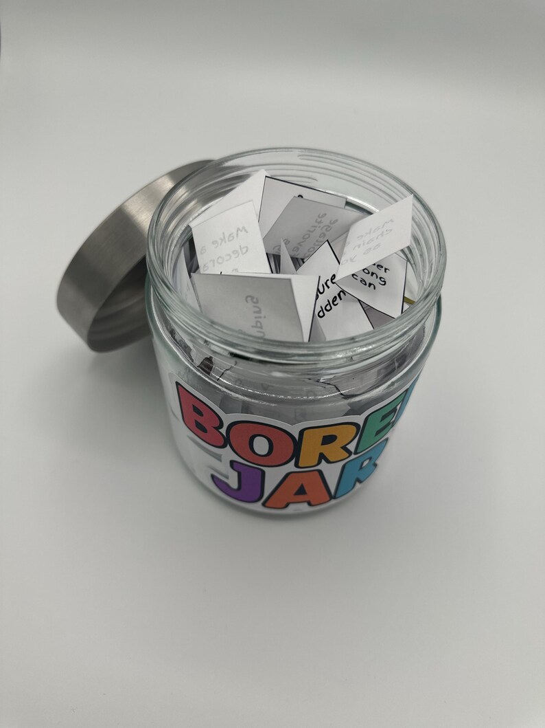 May include: A clear glass jar with a silver lid, filled with small white paper slips. The jar has a colorful label that reads "BORED JAR". The jar is sitting on a white surface.