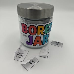 May include: A glass jar with a silver lid, labelled "BORED JAR" in colourful letters. Small folded white papers with handwritten text are scattered around the jar. The jar is filled with similar folded papers.
