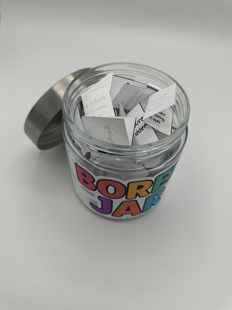 May include: A clear glass jar with a silver lid, filled with small white paper slips. The jar has a colorful label that reads "BORED JAR". The jar is sitting on a white surface.