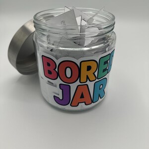 May include: A glass jar with a silver lid, filled with folded white paper slips. The jar features a colourful sticker that reads "BORED JAR" in a playful font. The lid is off to the side.