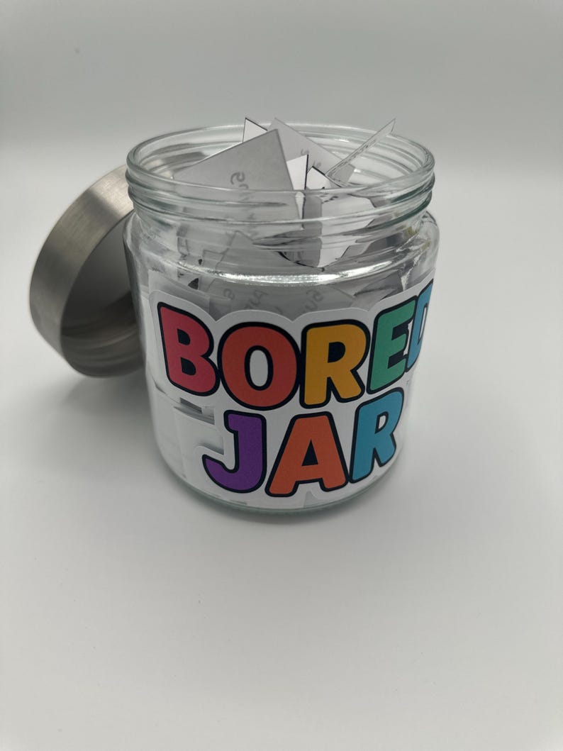 May include: A glass jar with a silver lid, filled with folded white paper slips. The jar features a colourful sticker that reads "BORED JAR" in a playful font. The lid is off to the side.