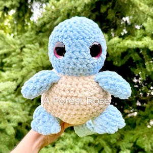 Peut inclure: Une peluche tortue au crochet faite à la main, de couleur bleu clair et beige. La tortue a de grands yeux bordés de rose et le texte "A Forest Rose" est visible en bas. La peluche est douce et câline.