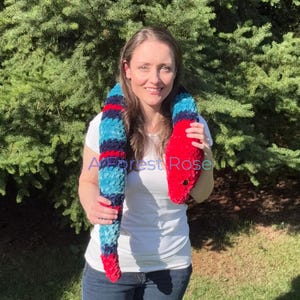 Puede incluir: Un juguete de serpiente de peluche hecho de hilo de terciopelo suave con rayas alternas en rojo brillante, azul marino oscuro y azul turquesa claro. La serpiente tiene una cabeza roja sólida con un pequeño ojo negro y el texto A Forest Rose.
