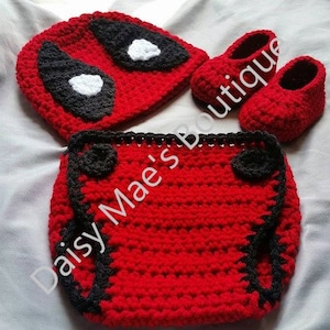 Könnte beinhalten: Ein gehäkeltes Baby-Outfit in Rot und Schwarz mit einem Deadpool-Design. Das Outfit beinhaltet eine Mütze, eine Windel-Abdeckung und Booties.