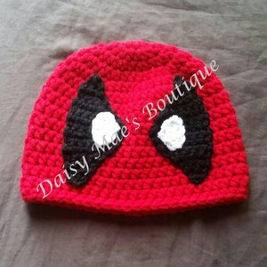 May include: Red crocheted hat with black and white eye mask design. The hat has the text "Daisy Mae's Boutique" on the side.