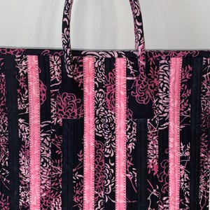 May include: A quilted tote bag with a floral pattern in black and pink. The bag features vertical stripes of pink and black fabric, with a matching handle. The bag is designed for carrying items.