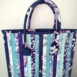 May include: A quilted tote bag with a floral pattern in shades of blue, purple, and white. The bag has a rectangular shape with two handles and a matching floral tag. The bag is made of fabric strips sewn together.