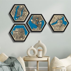 Puede incluir: Cuatro piezas de decoración de pared hexagonal de madera con marcos negros, cada una con un mapa detallado de una ciudad diferente. Los mapas están impresos en blanco y negro con características de agua azules. Las ciudades son Nueva York, Boston, San Francisco y Seattle.