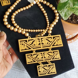 May include: A gold necklace with a layered pendant featuring a geometric design. The necklace is made of gold-toned beads and has a matching pair of earrings with a similar geometric design.
