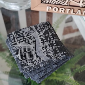 May include: A set of four black slate coasters featuring a laser-etched map of Portland, Oregon. The coasters are stacked on top of each other, with the top coaster showing the city's downtown area, including the Moda Center, Old Town, and the Pearl District. The text "Portland, OR" is visible at the bottom of the coaster.
