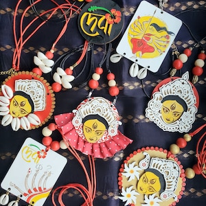 May include: A collection of six necklaces featuring colorful wooden pendants with images of the Hindu goddess Durga. The pendants are decorated with intricate details and are strung on red and black cords. The necklaces are adorned with white seashells and red and white beads.