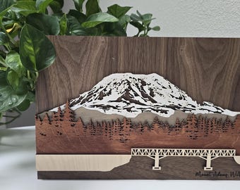 May include: Wooden wall art depicting a layered mountain landscape. The artwork shows a snow-covered mountain, trees, and a bridge, with the text "Mount Adams, WA" at the bottom. The piece is crafted from various shades of wood.