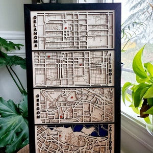 May include: A black framed wooden wall art piece with four laser-cut maps of different cities. The maps are labeled "Oklahoma", "Illinois", "Oregon", and "California".