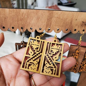 May include: A pair of light brown wooden earrings with a rectangular shape and intricate cut-out designs. The earrings are hanging from gold-colored hooks. Other earrings are visible in the background, displayed on a wooden rack.