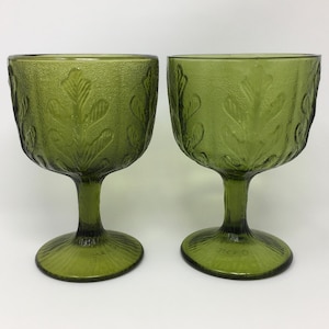 May include: Two green-tinted glass goblets. Each goblet features a textured leaf design on the bowl and stem. The goblets have a classic, vintage aesthetic, suitable for serving beverages. The goblets are made of glass.