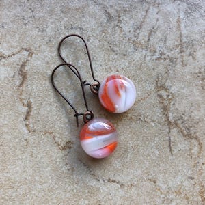 Puede incluir: Pendientes hechos a mano con cuentas de vidrio veteado en blanco, naranja y rosa. Las cuentas cuelgan de ganchos para las orejas de color bronce oscuro. Un accesorio único y artístico. Una adición elegante a cualquier colección de joyas.