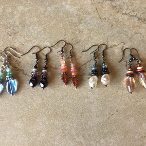 May include: Five pairs of dangle earrings featuring various bead combinations. The set includes blue teardrop beads, dark purple faceted beads, pink leaf-shaped beads, white irregular pearls, and clear amber-toned glass beads on metal hooks.