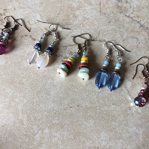 May include: Five pairs of dangle earrings featuring various bead designs. The first pair has purple glass beads, the second has translucent white stones, the third has multi-colored flat discs, the fourth has blue glass, and the fifth has dark purple beads.