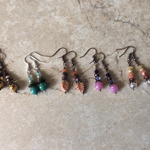 May include: Five pairs of dangle earrings featuring various bead designs. The earrings include spotted brown beads, turquoise spheres, carved leaf-shaped beads, round pink beads, and faceted clear and peach-colored beads, all suspended from metal hooks.