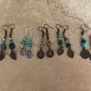 May include: Five pairs of dangle earrings featuring various beads in shades of blue, green, and brown, accented with metal charms including leaves, flowers, and circular medallions, all suspended from silver or copper-toned metal ear wires.