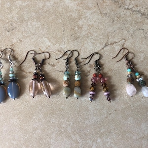 May include: Five pairs of dangle earrings featuring various bead combinations. The set includes grey teardrop beads, clear pink glass, multi-colored stone beads, faceted purple and brown crystals, and light pink stones with turquoise accents on metal hooks.