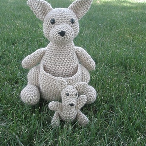 May include: A beige crocheted kangaroo with a smaller beige crocheted joey in its pouch. Both kangaroos have black eyes and are sitting on a green grassy surface.
