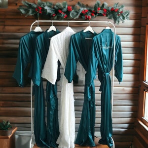 Puede incluir: Cinco batas colgadas en un estante, cuatro son azul turquesa y una es blanca. Las batas turquesa tienen la palabra "Bridesmaid" bordada en una de ellas. Las batas están colgadas frente a una pared de madera con una guirnalda de vegetación y bayas rojas.