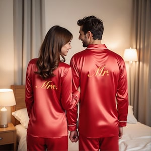 以下が含まれることがあります： お揃いの赤いサテンパジャマセット。長袖シャツは白いトリムと「Mrs.」と「Mr.」と金色の刺繍が施されています。二人は寝室で抱き合っています。パジャマはロマンチックなギフトのアイデアです。