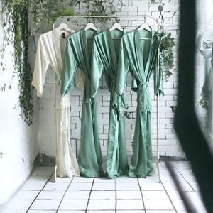 May include: Three green robes and one cream-colored robe hang on a metal rack against a white brick wall. The robes are made of a silky material and have tie closures. Greenery hangs from the top of the wall and the rack.