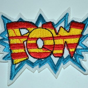 Può includere: Una toppa ricamata bianca con una grafica "POW" blu, rossa e gialla. Le lettere sono contornate di nero e hanno un motivo a righe.