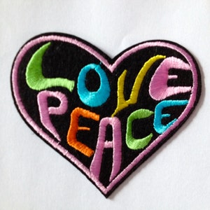 May include: An embroidered heart-shaped patch with the words "LOVE PEACE". The patch has a black background, a pink border, and colourful lettering in green, blue, orange, and yellow. Ideal for clothing or accessories.