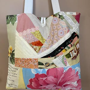 May include: A patchwork tote bag with white straps. The bag is made from various fabric patterns, including floral and geometric designs. The bottom of the bag features a large pink flower. The words "FAST COLOURS" and "ALL LINEN" are visible.