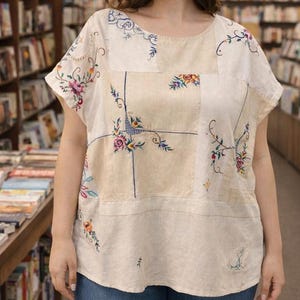 May include: Cream patchwork top with floral embroidery in pink, blue, and yellow. The short-sleeved shirt has a round neckline and is worn with blue jeans. The embroidery is detailed, giving a vintage look.