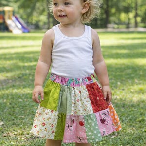 May include: A white sleeveless vest top paired with a colourful patchwork skirt featuring various patterns including floral prints, polka dots, and cherry motifs in shades of green, pink, red, and orange with an elasticated waistband.
