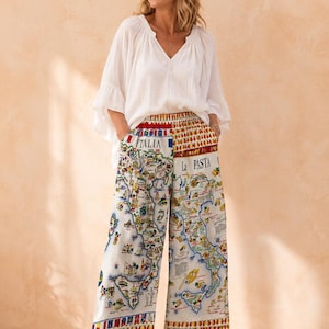 May include: A white V-neck blouse with loose, ruffled sleeves paired with wide-leg trousers featuring a colourful map print of Italy. The trousers display the text "ITALIA" and "La PASTA" with various illustrations of regional landmarks and food items.