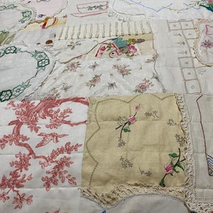 Peut inclure: Une courtepointe patchwork composée de divers morceaux de tissu, dont des imprimés floraux, des broderies et des bordures en dentelle. La courtepointe présente un mélange de couleurs, notamment du rouge, du rose et de la crème, avec les mots "Good Morning" brodés sur une section.