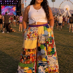 May include: A person wearing a white vest top and patchwork trousers with a variety of colourful patterns. The trousers have a wide leg design. The background shows a festival setting with a stage and a Ferris wheel.
