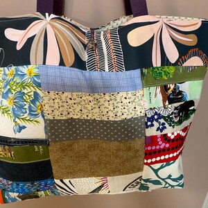 May include: A patchwork tote bag featuring a variety of fabric patterns including floral prints, blue gingham, and solid textures. The bag has two dark purple woven straps and a small silver-toned metal charm attached to the top centre opening.
