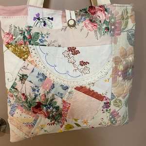 May include: A patchwork tote bag constructed from various floral and patterned fabric scraps in shades of pink, white, and green. The bag features a central embroidered semi-circular lace-trimmed pocket, a button closure, and two cream-coloured straps.
