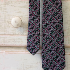 May include: Two patterned neckties in black, green and burgundy. The ties have a geometric design and are hanging on a white dresser.