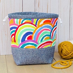 May include: A drawstring project bag with a rainbow pattern and a denim bottom. The bag is filled with yellow yarn.