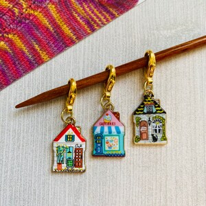 May include: Three gold-toned metal stitch markers shaped like houses with different storefront designs. The houses are decorated with colourful details and text, including "Cafe", "Cupcakes", and a black cat.
