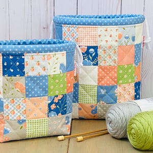 May include: Two quilted fabric storage baskets featuring a patchwork design of blue, peach, green, and white squares with floral, gingham, and peach motifs. Each basket has a blue patterned top border, white drawstring cords, and quilted stitching.