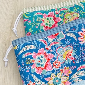Peut inclure: Deux sacs en tissu à cordon avec des motifs floraux. Le sac du haut est turquoise avec des fleurs blanches et roses. Le sac du bas est bleu avec des fleurs roses, blanches et jaunes.